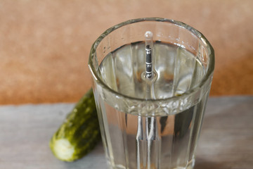 Glass of homemade moonshine with alcoholometer and pickled cucumber on the wooden background. Closeup, selective focus