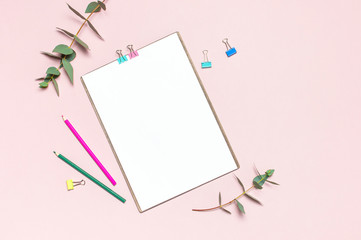 Flat lay clipboard with blank paper, colour pencils, clips, eucalyptus branches on pink background. Top view female office workspace minimal mock up template concept copy space Feminine floral desktop