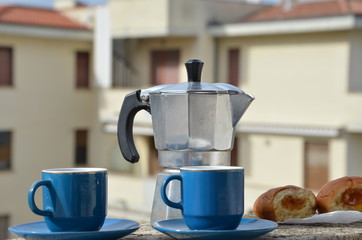 Coffee maker moka pot with roasted coffee beans. Space for text, horizontal. Breakfast concept. Pot and two blue cups of coffee on the balcony with a croissant. On the background of the city.