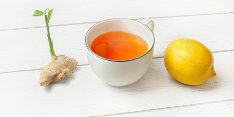 Hot amber tea in porcelain cup, whole lemon and ginger root with green sprout next to it on white boards.