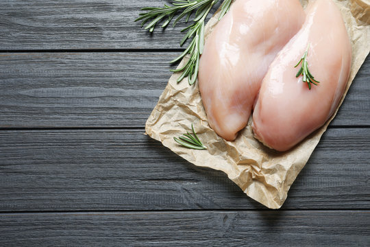 Parchment with raw chicken breasts and rosemary on wooden background, top view. Space for text