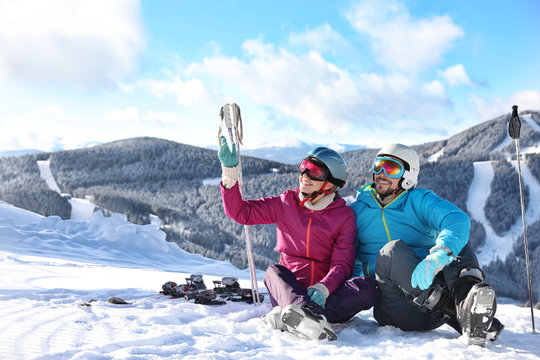 Happy couple with ski equipment sitting on snowy hill in mountains, space for text. Winter vacation