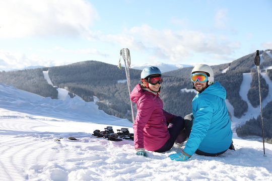 Happy couple with ski equipment sitting on snowy hill in mountains, space for text. Winter vacation