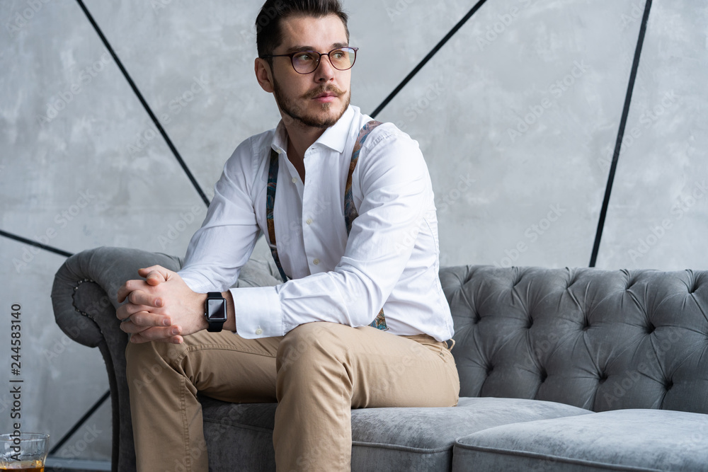 Wall mural enjoying free time. thoughtful young man in full suit looking away while sitting on the sofa at home