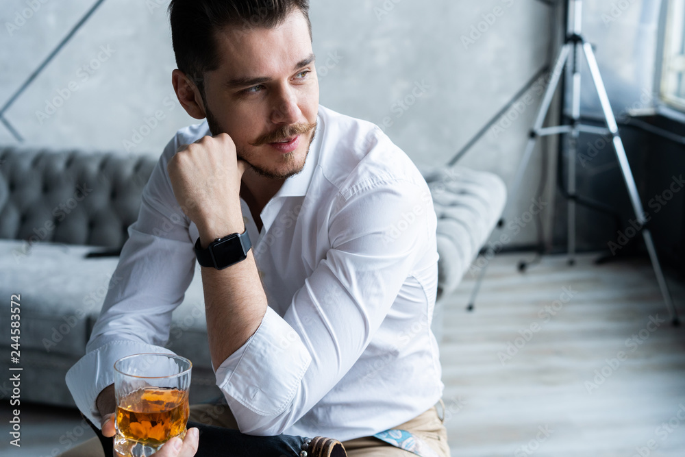 Wall mural Feeling lonely. Stylishly dressed young man holding a glass while sitting on on the stool.