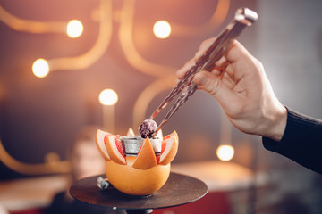 Fototapeta na wymiar Waiter straightens coal Hookah shisha with tobacco on orange fruit in nightclub or bar
