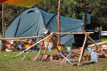 Tourist life - camp tent with a crossbar over the fire on the green grass field on the forest background on summer day - sports tourism, camping, scouting