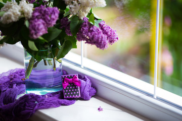 Purple gift box and tender bouquet of beautiful lilac in glass vase near window in daylight.