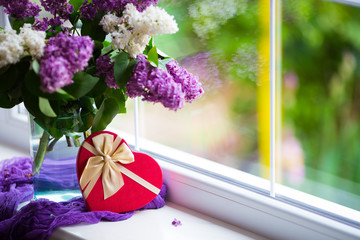 Heart shaped gift box and tender bouquet of beautiful lilac in glass vase near window in daylight.