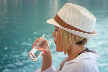 Caucasian blonde girl drinks cold water from a sweaty glass on the background of the turquoise sea