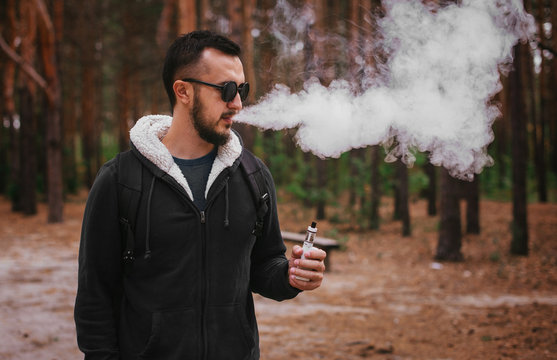 man vaping electronic cigarette in nature. man with beard in glasses