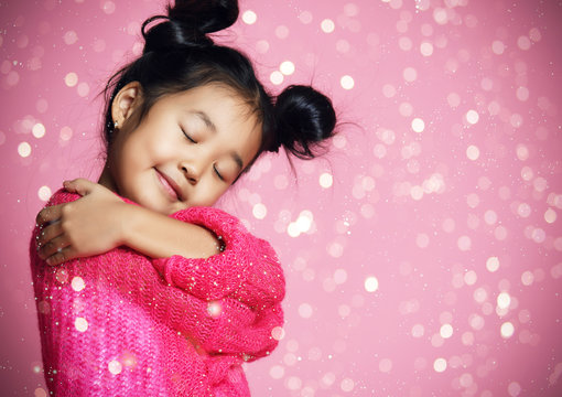 Asian kid girl with closed eyes in pink sweater hug herself and dream. golden spangles