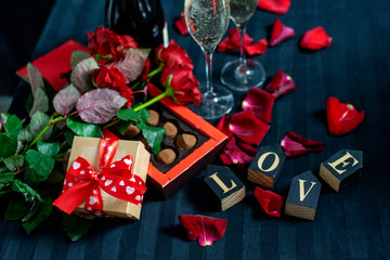 two glasses of champagne, red roses, petals, gift box with red ribbon, chocolates and wooden love words on a black background.