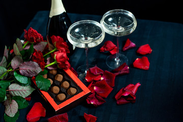 two glasses of champagne, red roses, petals and chocolates on a black background