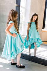 The lovely little girl posing in a beautiful blue dress is posing looking at the mirror