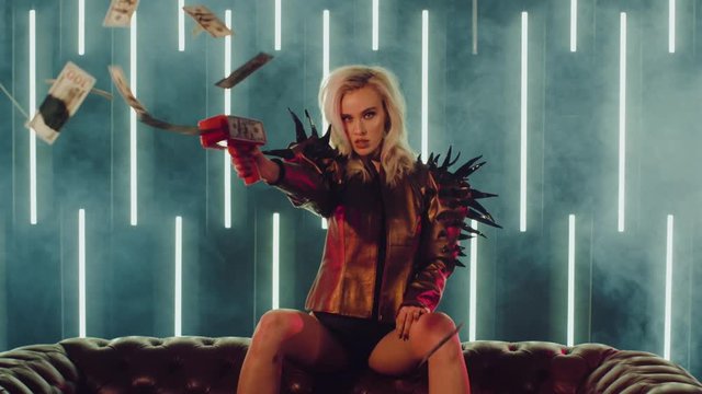 Sexual young girl is firing from gun with dollars in nightclub. She is pulling the trigger and banknotes are flying. Shoot with RED RAVEN camera