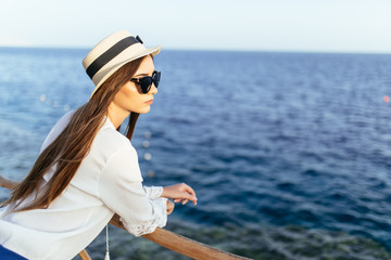 Young prety woman in straw hat and sunglasses on pier in stylish elegant clothes poses enjoying amazing view.