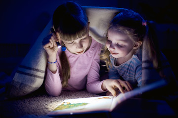 two sisters reading a book with a flashlight in a dark room at night