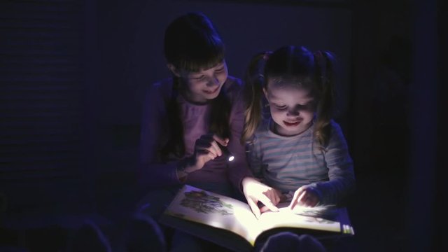 two sisters reading a book with a flashlight in a dark room at night