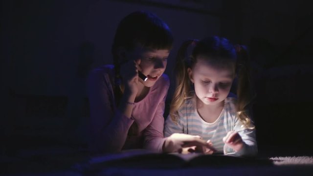 two sisters reading a book with a flashlight in a dark room at night