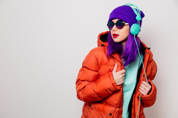 Beautiful young girl with purple hair and in orange jacket listen music in headphones on white...