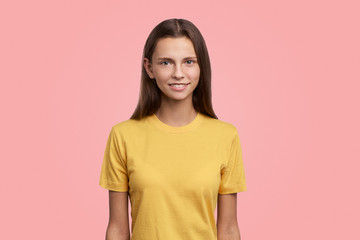 Obraz premium Photo of pretty teen girl has serious look at camera, long hair, has gentle smile, shows her natural beauty. Teenager poses against pink background with copy space for advertisement