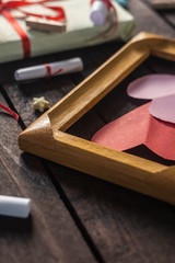 Flat lay romantic background. Giftbox, rolled wish papers, and wooden frame with paper hearts on dark wood board.