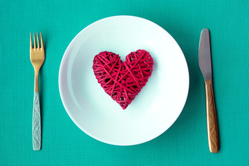 Festive table setting for Valentine's Day. Red heart on a white plate with a knife and fork on a greene background.