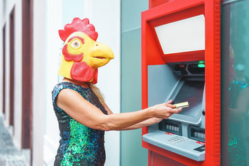 Mature woman wearing chicken mask withdraw money from bank cash machine with debit card - Surreal...