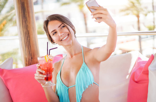 Young pretty woman taking a selfie with mobile smart phone in a beach club - Attractive girl having fun drinking a tropical cocktail and posting photos on her social networs during her vacation