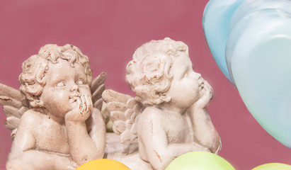 Two chipped cherub statues with chins on hands surrounded by pastel hearts isolated on pink background for Valentine's Day