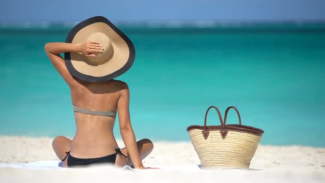 Beach woman looking at sea while sunbathing on beach holding sunhat from wind. Woman is in bikini during vacation. Sexy lady is enjoying summertime at beach. Summer Vacation Concept