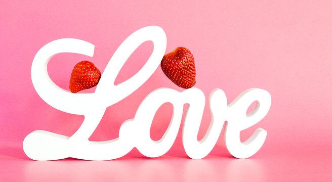 Valentine's Day-white wooden letters spelling the word LOVE with two ripe red strawberries leaning against the L isolated on a pink background