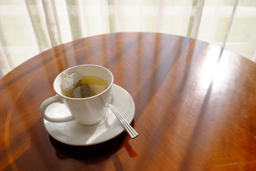 Full glass of instant tea with saucer and metal spoon on round glossy wooden table with sunlight through the curtain of window
