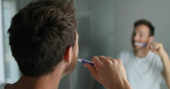Man brushing his teeth getting ready in the morning doing hygiene routine looking in mirror of home bathroom using toothbrush in for clean dental oral care.