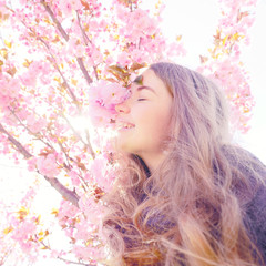 Fototapeta premium Girl smiling and standing near sakura flowers, defocused. Girl with long hair outdoor, cherry blossom on background. Perfume and fragrance concept. Woman enjoy aroma of sakura on spring day.