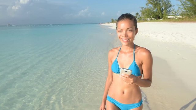 Beach vacation girl using mobile phone app textng sms or browsing on online social media during summer travel holiday. Woman walking in bikini on sand lying on towel wearing sun hat. 59.94 FPS