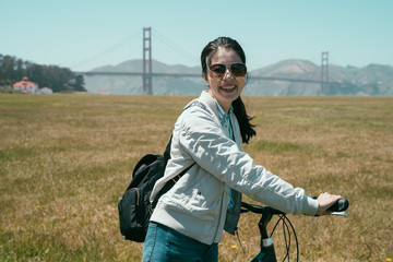 young female tourist wearing sunglasses walking cheerful with bicycle on the field under sunshine in summer. golden state bridge famous attraction in background. girl traveler love hobby sport cycle.