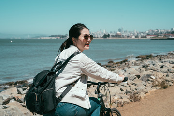 Fototapeta premium cheerful lady traveler riding bicycle along the coast on the sand on sunny day. young asian woman wearing sunglasses carrying backpack cycling sightseeing outdoor. girl backpacker travel Marin county