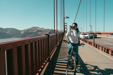 Golden gate bridge biking lady sightseeing in San Francisco USA. Young female tourist on bike tour enjoying the view stop riding bicycle taking picture shooting photographing on sunny day in summer.