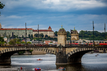 Fototapeta premium PRAGUE, CZECH REPUBLIC - AUGUST 28, 2015: Pedal boats sail and pass under the medieval stone Chec Bridge on Vltava River, Prague, Czech Republic in calm summer evening with people walking on top