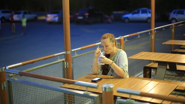 young single woman sitting in the evening in a summer cafe on the street , listening to music on smartphone and have dessert.