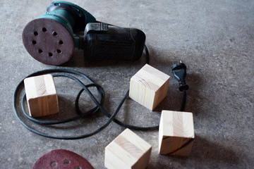 Dusty electrical orbital sander with cable, sanding disc and natural raw wooden cubes on grey grunge table. Carpentry equipment and making wooden toys diy concept.