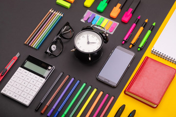 Flat lay composition of business desk with alarm clock, smartphone, notebook, stickers, and colored pens on colorful black and yellow background