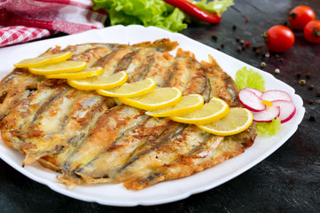 Fried capelin with lemon on a white plate on a black wooden background. A dish of small sea fish. Close up