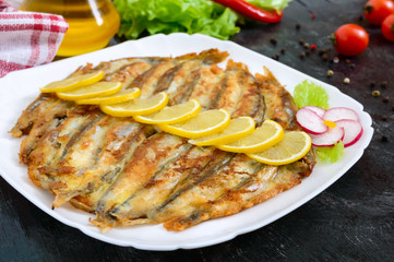 Fried capelin with lemon on a white plate on a black wooden background. A dish of small sea fish. Close up
