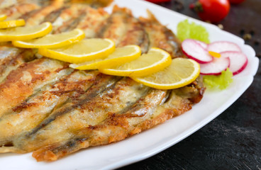 Fried capelin with lemon on a white plate on a black wooden background. A dish of small sea fish. Close up