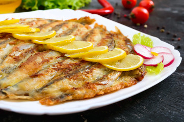 Fried capelin with lemon on a white plate on a black wooden background. A dish of small sea fish. Close up