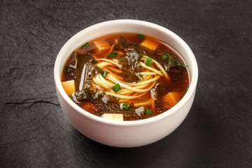A photo of a bowl of miso shiru soup with tofu, scallions, noodles, and wakame seaweed, on a black background