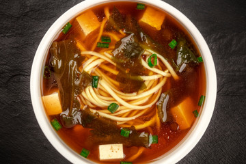 A closeup photo of a bowl of miso shiru soup with tofu, scallions, noodles, and wakame seaweed, shot from the top on a black background